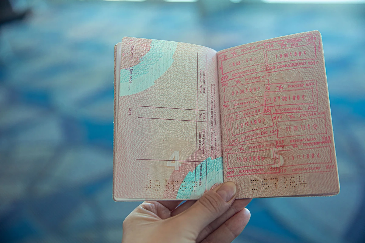 A hand holding an open passport whose page is covered in red entry and exit stamps, against a blurred blue airport background